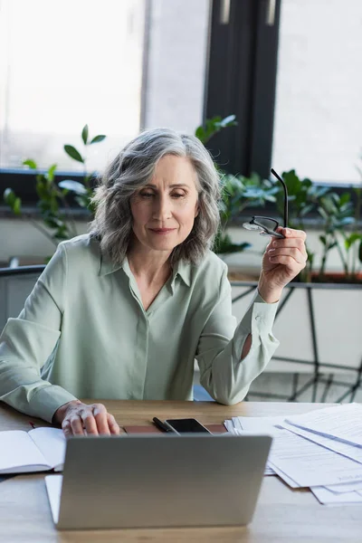 Middle aged businesswoman holding eyeglasses near gadgets, papers and notebook in office — Stock Photo