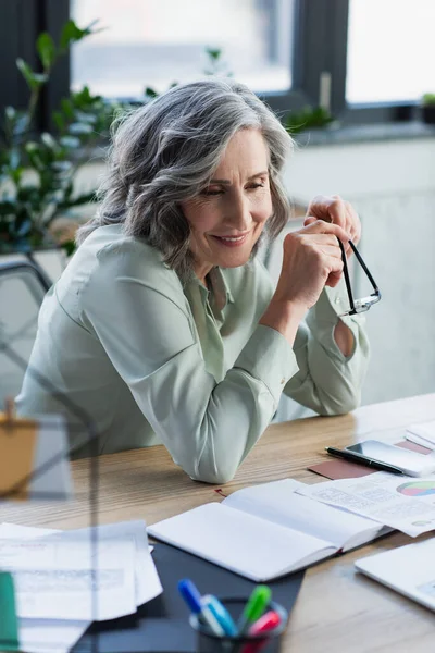 Cheerful businesswoman holding eyeglasses near documents and gadgets on table in office — Stock Photo