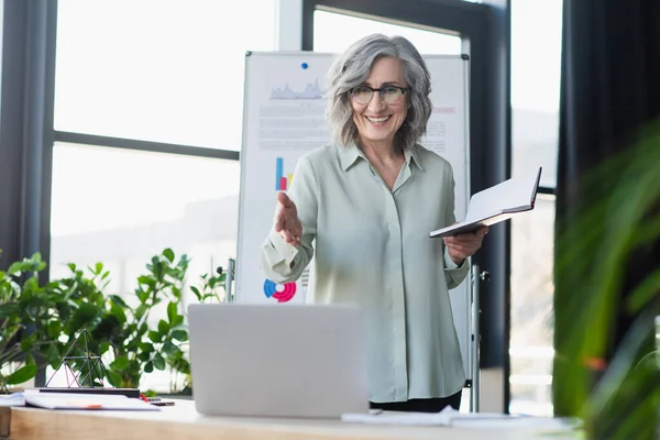 Happy businesswoman holding notebook during video call on laptop in office — Stock Photo