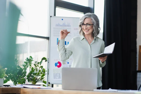 Sorrindo mulher de negócios madura apontando para flipchart e segurando notebook perto de laptop no escritório — Fotografia de Stock