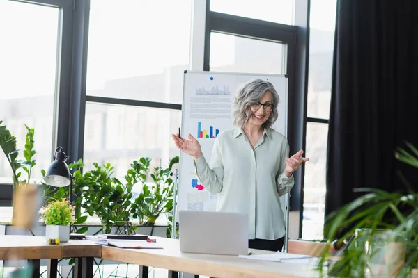 Glückliche reife Geschäftsfrau mit Videoanruf auf Laptop in der Nähe von Flipchart im Büro — Stockfoto