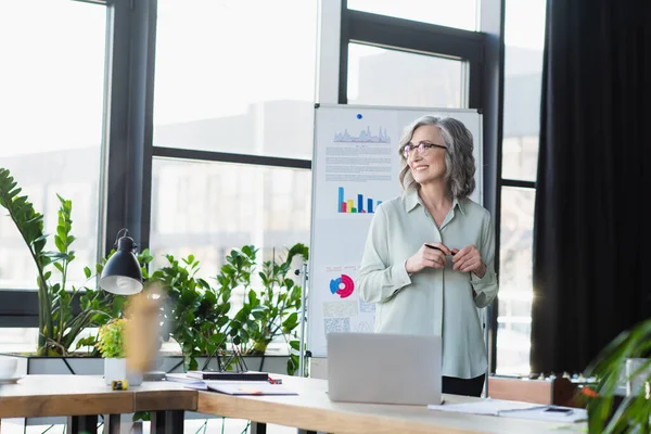 Серая деловая женщина в очках держит ручку и улыбается возле флип-чарта и рабочего стола в офисе — стоковое фото