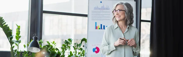 Alegre mujer de negocios sosteniendo pluma cerca de rotafolio y lámpara en la oficina, pancarta - foto de stock