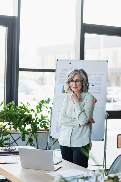 Businesswoman in eyeglasses looking at laptop near flip chart in office — Stock Photo