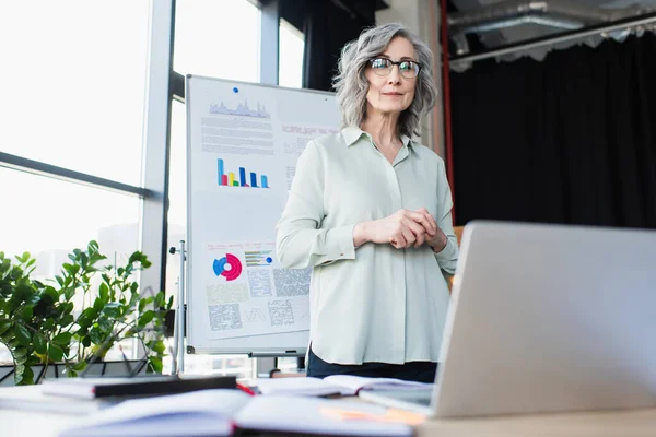 Middle aged businesswoman in eyeglasses looking at laptop near flip chart in office — Stock Photo