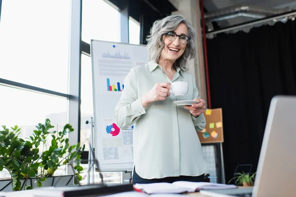 Smiling businesswoman holding coffee cup and looking at laptop in office — Stock Photo