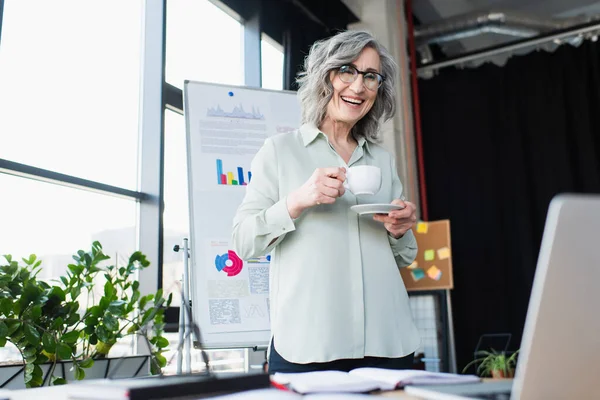 Donna d'affari sorridente che tiene la tazza di caffè e guarda il computer portatile in ufficio — Foto stock