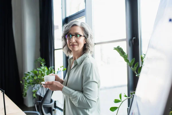 Mujer de negocios de pelo gris sosteniendo café cerca de rotafolio en la oficina - foto de stock