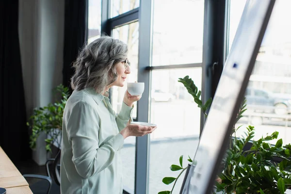 Vue latérale d'une femme d'affaires mature tenant une tasse de café près d'un tableau à feuilles brouillées au bureau — Photo de stock