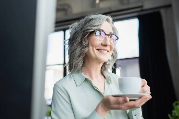 Joyeux gris femme d'affaires aux lunettes tenant tasse et soucoupe au bureau — Photo de stock