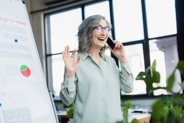 Mature businesswoman talking on cellphone near flip chart in office — Stock Photo