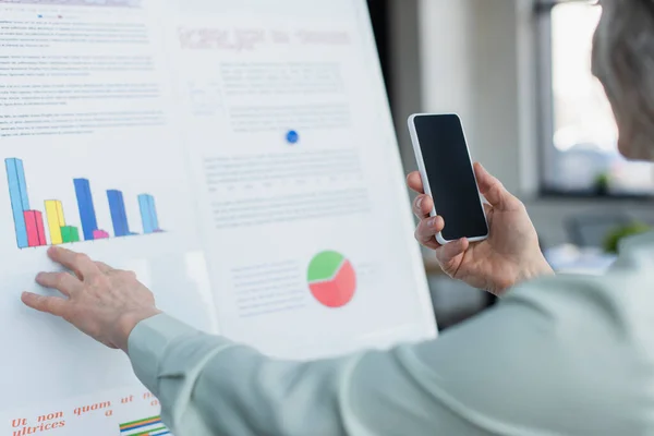 Cropped view of mature businesswoman taking photo on cellphone near flip chart with graphs — Stock Photo