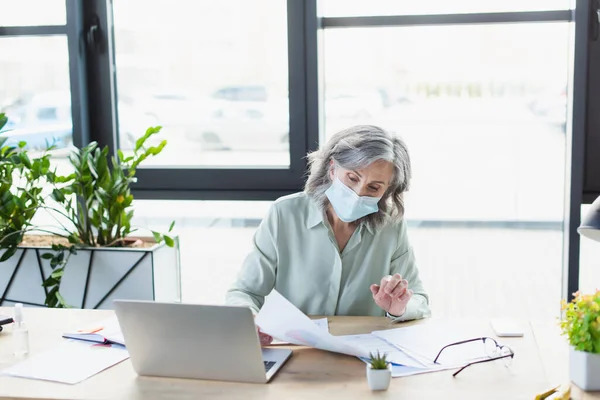 Зріла бізнес-леді в медичній масці, що працює з документами біля ноутбука в офісі — стокове фото