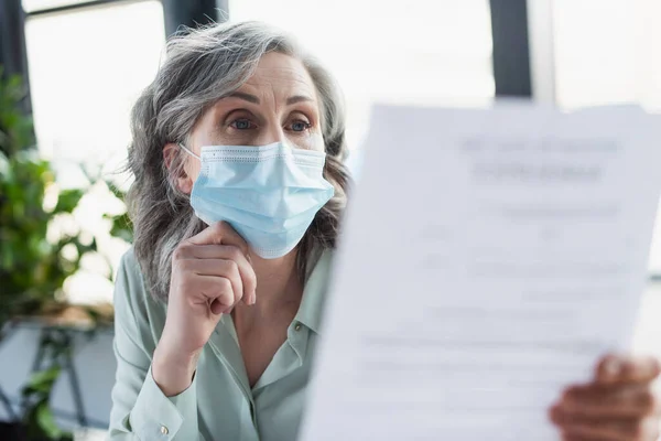 Donna d'affari dai capelli grigi in maschera medica guardando il documento sfocato in ufficio — Foto stock