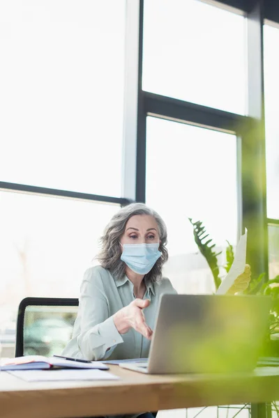 Femme d'affaires dans le masque médical ayant appel vidéo sur ordinateur portable dans le bureau — Photo de stock