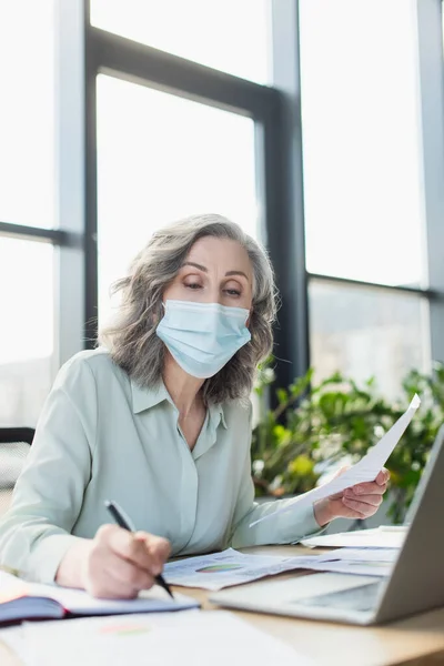 Donna d'affari dai capelli grigi in maschera medica con carta e scrittura su notebook vicino al laptop in ufficio — Foto stock