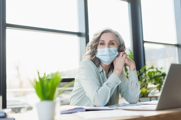 Reife Geschäftsfrau in medizinischer Maske schaut in der Nähe von Laptop im Büro in die Kamera — Stockfoto