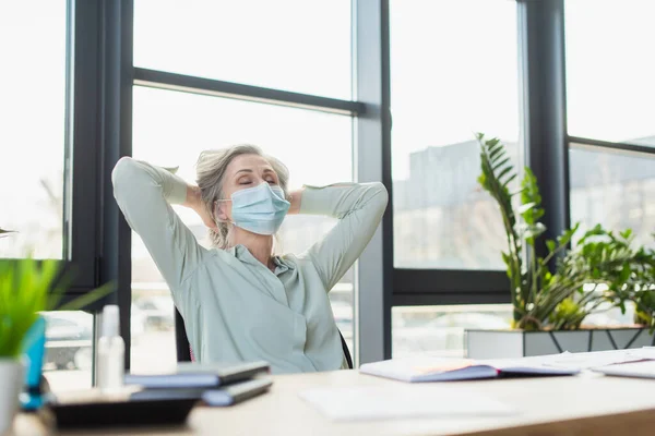 Donna d'affari matura in maschera medica seduta con gli occhi chiusi vicino notebook in ufficio — Foto stock