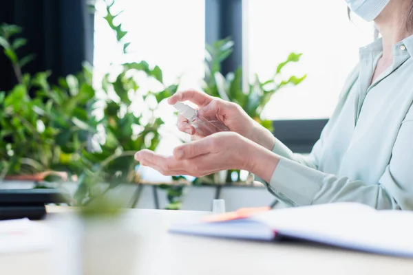 Cropped view of businesswoman in medical mask using hand sanitizer near notebook in office — Stock Photo