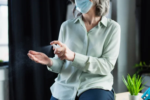 Vista ritagliata di donna d'affari dai capelli grigi in maschera protettiva spruzzando disinfettante mano in ufficio — Foto stock