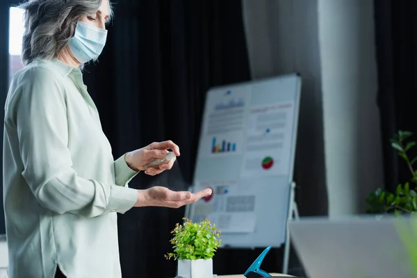 Empresaria de mediana edad en máscara médica rociando desinfectante de manos en la oficina - foto de stock