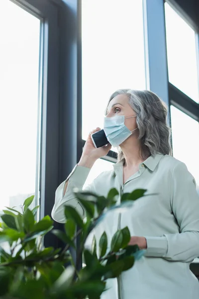 Madura mujer de negocios en máscara médica hablando en el teléfono inteligente cerca de la planta y ventana en la oficina - foto de stock