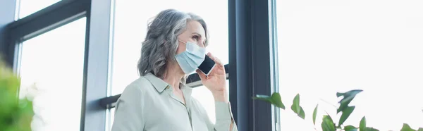 Mujer de negocios de pelo gris en máscara médica hablando en el teléfono inteligente cerca de la ventana en la oficina, pancarta - foto de stock