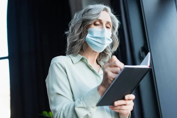 Visão de ângulo baixo de empresária de cabelos grisalhos em máscara médica segurando notebook no escritório — Fotografia de Stock