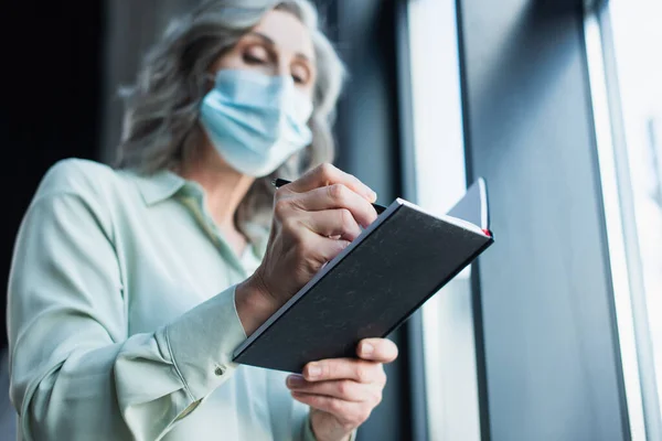 Vista de ángulo bajo de la mujer de negocios borrosa en la escritura de la máscara médica en el cuaderno en la oficina - foto de stock