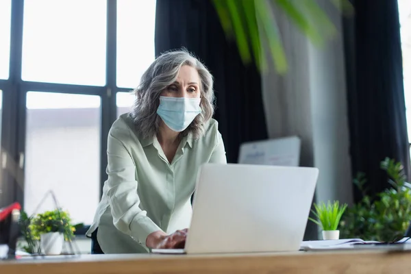 Donna d'affari di mezza età in maschera medica utilizzando laptop in ufficio — Foto stock