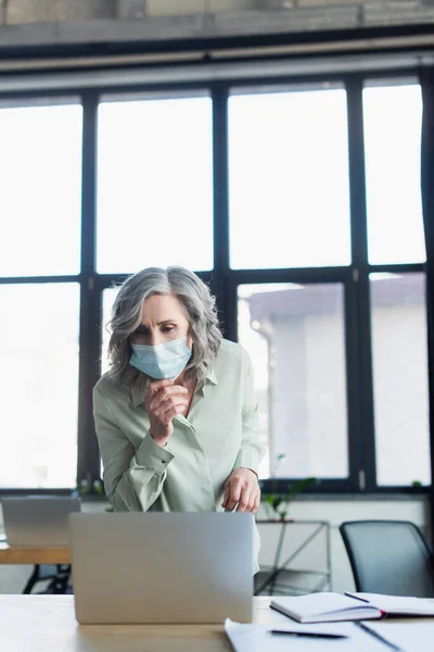 Mature businesswoman in protective mask looking at laptop near notebooks in office — Stock Photo