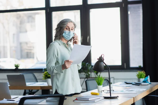 Зрелая деловая женщина в медицинской маске разговаривает на смартфоне и держит документ в офисе — стоковое фото