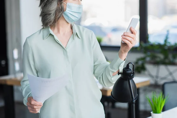 Обрезанный вид седой предпринимательницы в медицинской маске, держащей бумагу и мобильный телефон в офисе — стоковое фото