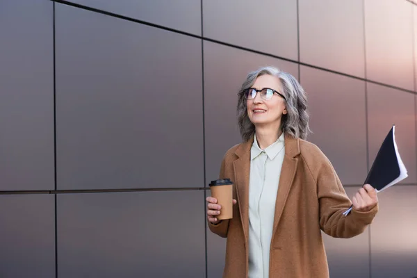 Heureuse femme d'affaires en manteau tenant du café pour aller et dossier en papier près du bâtiment à l'extérieur — Photo de stock