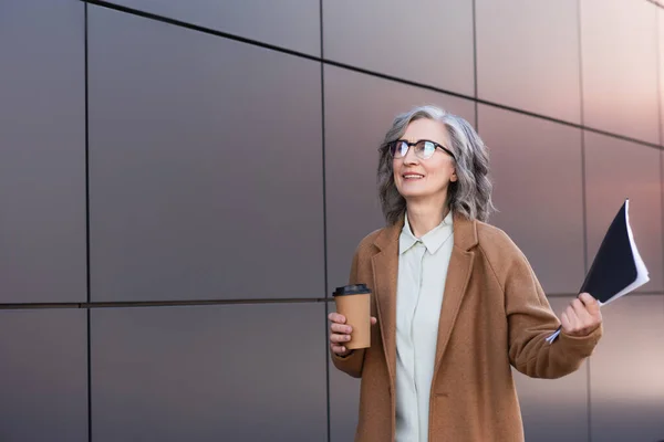 Happy businesswoman in coat holding coffee to go and paper folder near building outdoors — Stock Photo