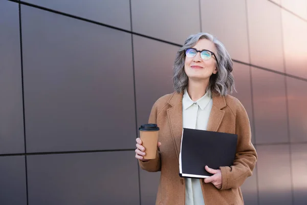 Grey haired businesswoman in eyeglasses and coat holding takeaway drink and paper folder outdoors — Stock Photo