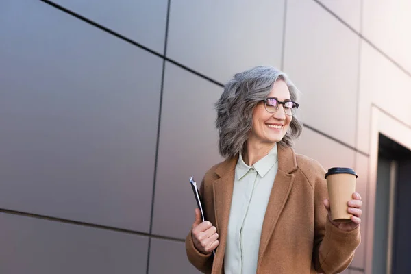Cheerful businesswoman in coat looking at paper cup while holding documents outdoors — Stock Photo