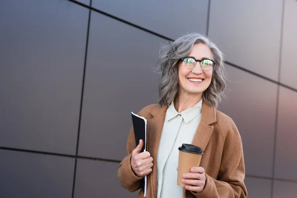 Деловая женщина средних лет в пальто и очках держит бумажную папку и кофе, чтобы выйти на улицу — стоковое фото