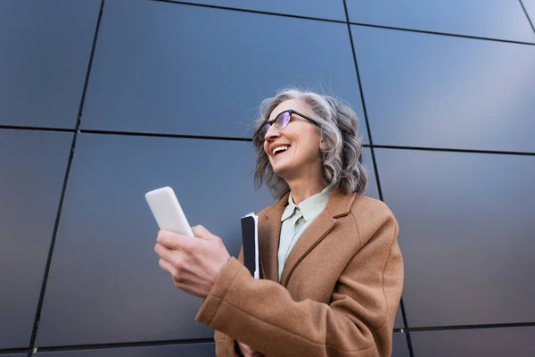 Vue en angle bas de femme d'affaires gaie dans le manteau tenant le téléphone portable et le dossier de papier près du bâtiment à l'extérieur — Photo de stock
