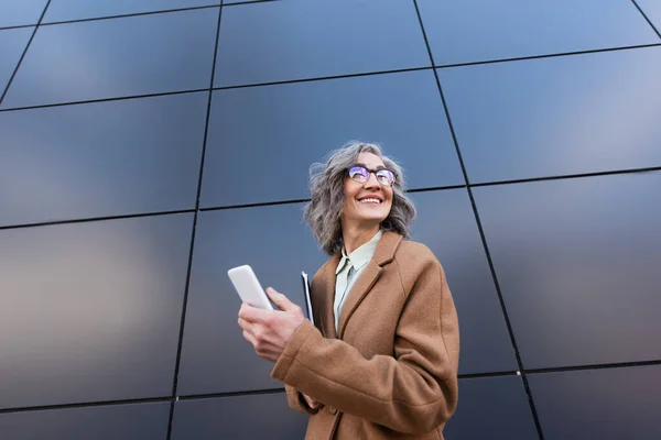 Vista de ángulo bajo de la mujer de negocios feliz en abrigo con teléfono inteligente y carpeta de papel cerca del edificio al aire libre - foto de stock