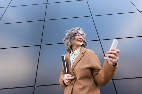 Vista de ángulo bajo de la alegre mujer de negocios madura con abrigo usando teléfono celular y sosteniendo la carpeta de papel cerca del edificio al aire libre - foto de stock