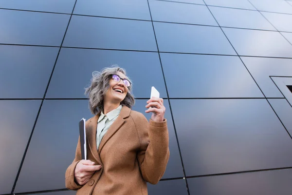 Low angle view of happy mature businesswoman in coat holding smartphone and paper folder near building outdoors — Stock Photo