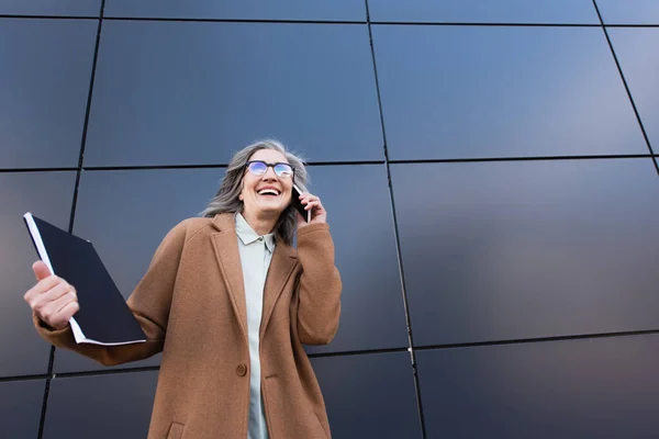 Donna d'affari sorridente in appendiabiti cartella di carta mentre parla su smartphone vicino edificio sulla strada urbana — Foto stock