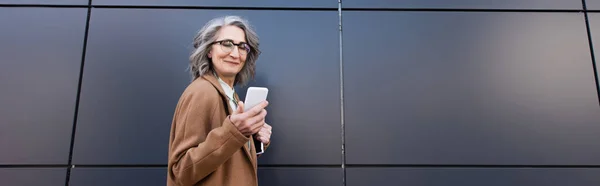 Empresária de meia idade em casaco e óculos segurando celular e pasta de papel ao ar livre, banner — Fotografia de Stock