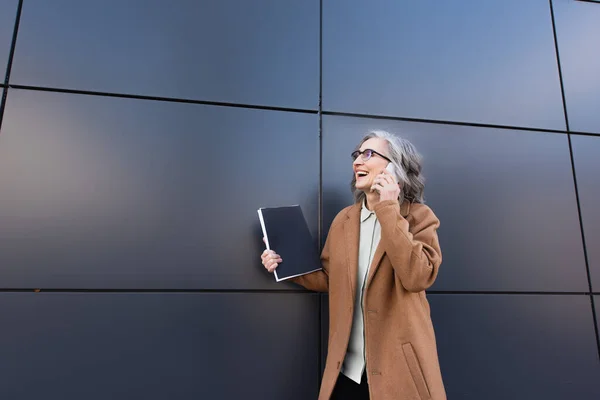 Mature businesswoman in coat talking on mobile phone and holding documents near building outdoors — Stock Photo