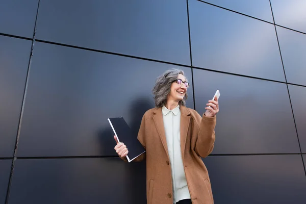 Щаслива бізнес-леді в пальто дивиться на смартфон і тримає паперову папку на відкритому повітрі — стокове фото