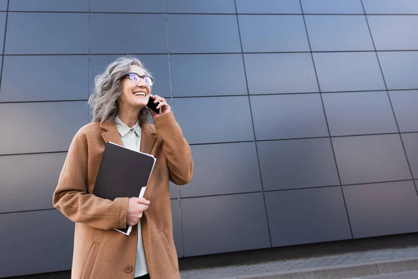 Веселая седовласая деловая женщина с газетами, разговаривающая по мобильному телефону на городской улице — стоковое фото