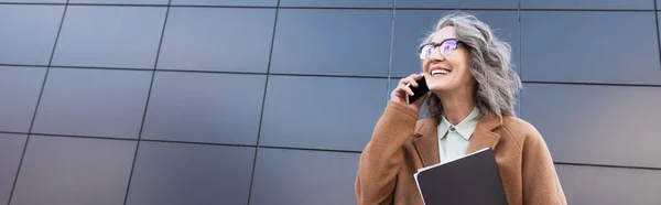 Positive businesswoman in coat holding documents and talking on mobile phone outdoors, banner — Stock Photo