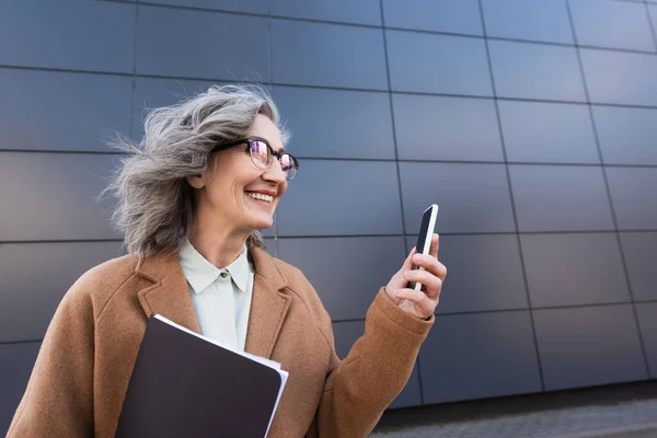 Mature businesswoman in eyeglasses holding cellphone and papers outdoors — Stock Photo