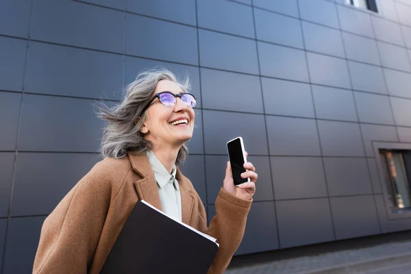 Happy businesswoman in coat holding paper folder and cellphone with blank screen on urban street — Stock Photo